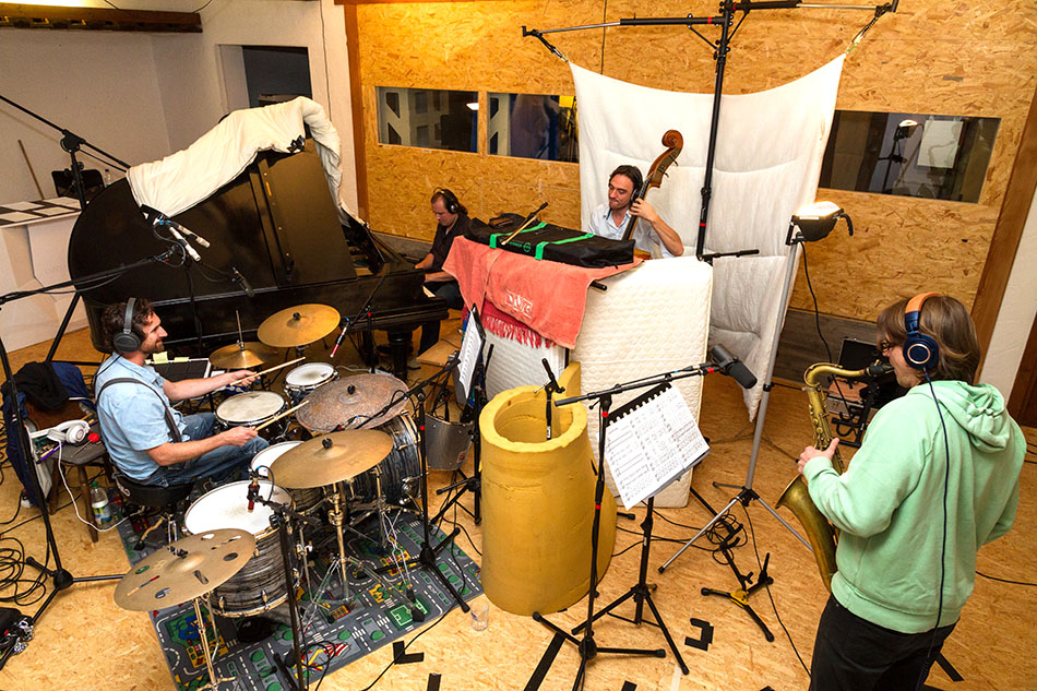 Full live-room recording setup: drums left foreground, piano centre-left background, upright bass centre-right background, saxophone right foreground